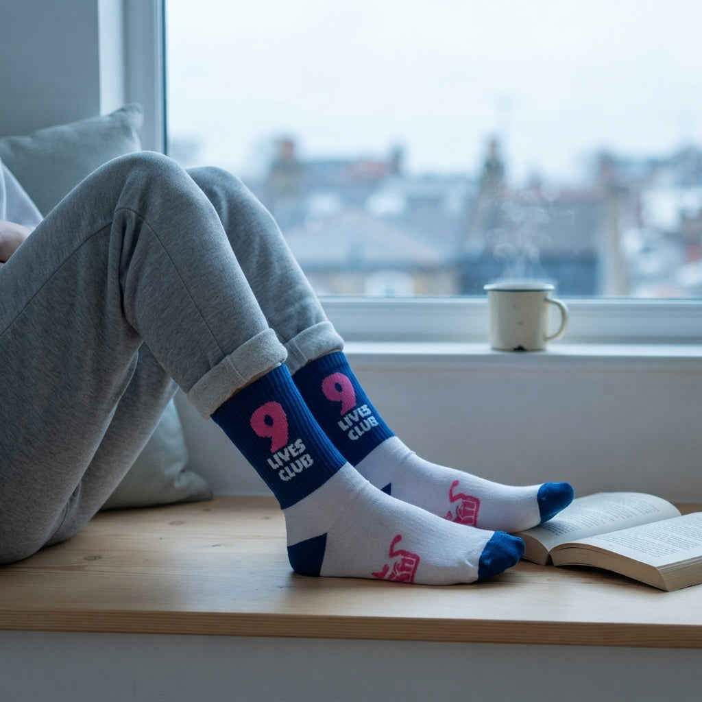 White crew socks with blue toes and heels, featuring a dark blue cuff with pink '9 LIVES CLUB' text, worn by a person sitting on a wooden window sill with an open book and a mug nearby.